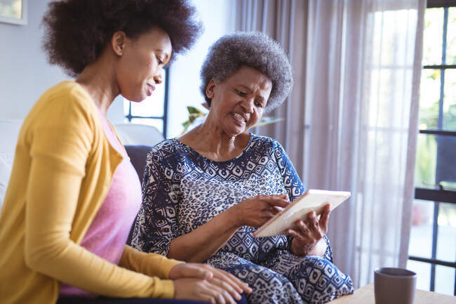 Femme âgée afro-américaine souriante avec fille adulte utilisant une tablette. temps en famille à la maison en utilisant la technologie ensemble. — Photo de stock