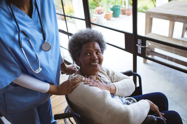 Médecin afro-américaine traitant patiente âgée souriante en fauteuil roulant à la maison. soins de santé et mode de vie pendant la pandémie de covide 19. — Photo de stock