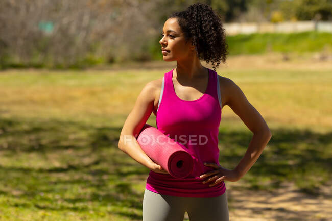 Relaxing biracial woman standing and holding yoga mat in countryside. healthy, active outdoor lifestyle and leisure time. — Stock Photo