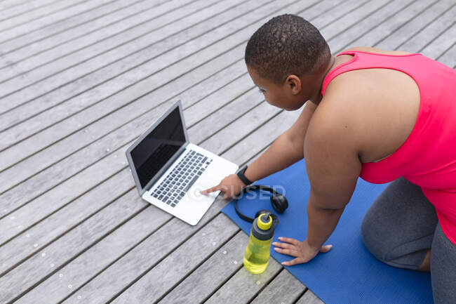 Afroamerikanische Plus-Size-Frau in Sportkleidung sitzt auf Matte und benutzt Laptop mit Kopierplatz. Fitness und gesunder, aktiver Lebensstil. — Stockfoto
