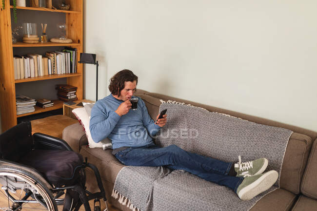 Кавказький інвалід п'є каву і користується смартфоном, який сидить вдома на дивані. інвалідність і гандикап — стокове фото