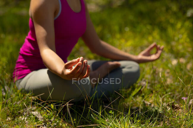 Seção intermediária de mulher biracial relaxante sentado com pernas cruzadas e meditando no campo. saudável, estilo de vida ao ar livre ativo e tempo de lazer. — Fotografia de Stock