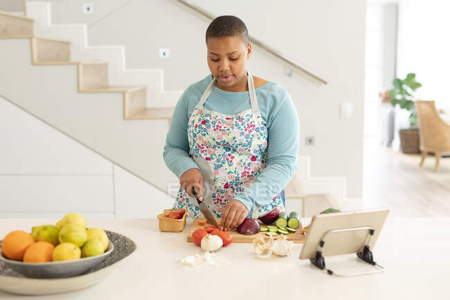 Afro-Américaine plus taille femme coupant des légumes, en utilisant une tablette dans la cuisine. mode de vie, cuisine et passer du temps à la maison. — Photo de stock