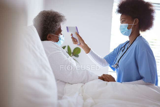 Médico afroamericano con mascarilla facial tomando la temperatura de la paciente anciana en casa. salud y estilo de vida durante la pandemia de covid 19. - foto de stock