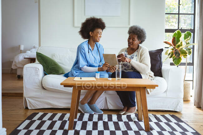 Улыбающаяся африканская американка-врач использует планшет со старшей пациенткой дома. здравоохранение и образ жизни во время пандемии ковида 19. — стоковое фото