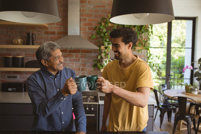 Sonriente hijo adulto birracial y padre mayor bebiendo café en la cocina. tiempo en familia en casa juntos. - foto de stock
