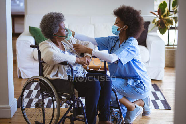 African american female doctor with face mask treating senior female patient on wheelchair at home. healthcare and lifestyle during covid 19 pandemic. — Stock Photo