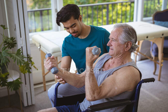 Fisioterapeuta birracial masculino sonriente tratando los brazos de un paciente masculino mayor en silla de ruedas en la clínica. atención médica de alto nivel y tratamiento de fisioterapia médica. - foto de stock