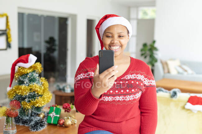 Heureuse femme afro-américaine plus taille dans le chapeau de Père Noël faire appel vidéo de Noël sur smartphone. Noël, fête et technologie de la communication. — Photo de stock