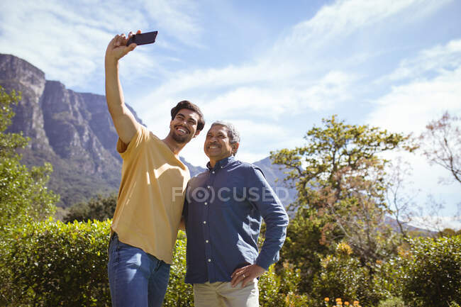 Sorrindo biracial filho adulto e pai sênior tomando selfie com smartphone no jardim. tempo de família em casa juntos. — Fotografia de Stock