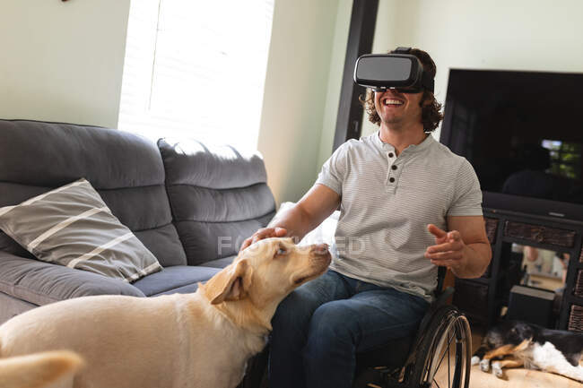 Uomo disabile caucasico con auricolare VR seduto sulla sedia a rotelle toccare il suo cane a casa. concetto di disabilità e handicap — Foto stock