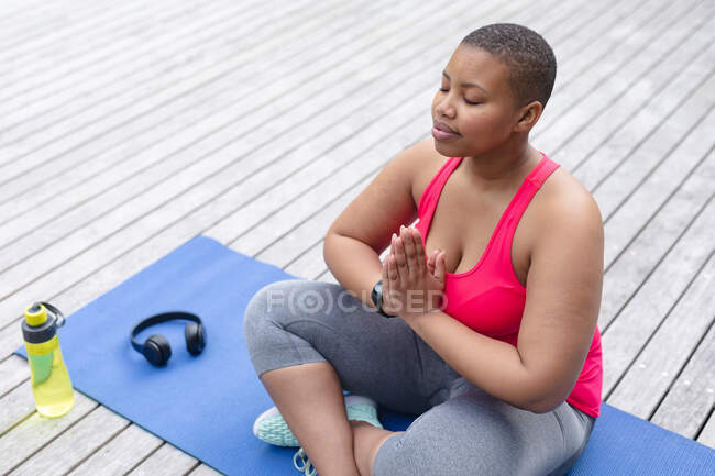Femme afro-américaine plus la taille en vêtements de sport assis sur le tapis et pratiquant le yoga. forme physique et mode de vie sain et actif. — Photo de stock