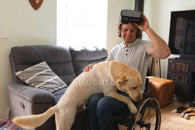 Uomo disabile caucasico con auricolare VR seduto sulla sedia a rotelle toccare il suo cane a casa. concetto di disabilità e handicap — Foto stock