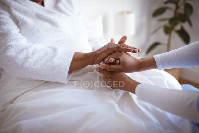 African american female doctor holding hands of senior female patient at home. healthcare and lifestyle during covid 19 pandemic. — Stock Photo