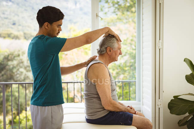 Fisioterapeuta masculino biracial tratando pescoço de paciente masculino sênior na clínica. cuidados de saúde seniores e tratamento de fisioterapia médica. — Fotografia de Stock