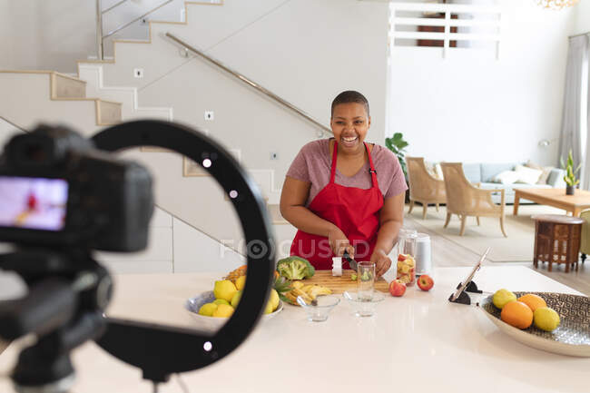 Feliz afroamericano más tamaño mujer cortando frutas, haciendo vlog en cocina. estilo de vida, cocinar y pasar tiempo en casa. - foto de stock