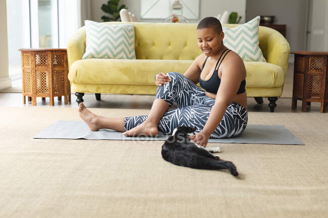 Щаслива афроамериканка плюс розмір жінка практикує йогу на килимку вдома з кішкою. фітнес і здоровий, активний спосіб життя . — стокове фото
