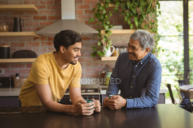 Sonriente hijo adulto birracial y padre mayor hablando y tomando café en la cocina. tiempo en familia en casa juntos. - foto de stock