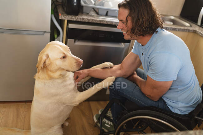 Kaukasischer Behinderter, der im Rollstuhl sitzt und zu Hause mit Hund spielt. Behinderten- und Behindertenkonzept — Stockfoto