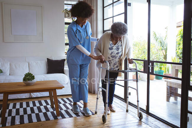 African american female doctor helping senior female patient with walking frame at home. healthcare and lifestyle during covid 19 pandemic. — Stock Photo