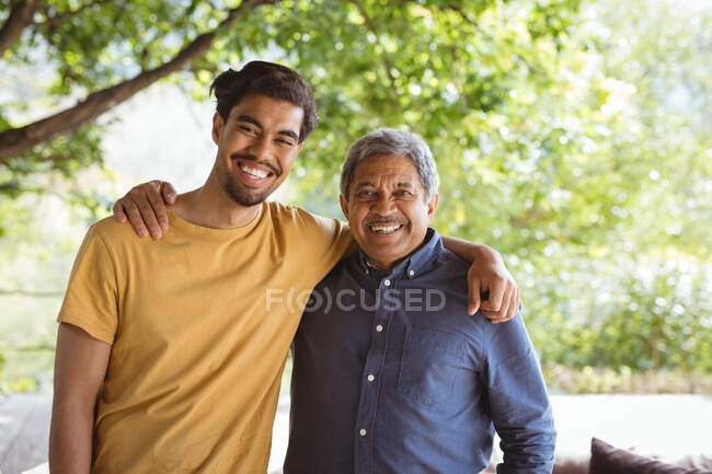 Portrait of smiling biracial adult son and senior father looking at camera and embracing. family time at home together. — Stock Photo