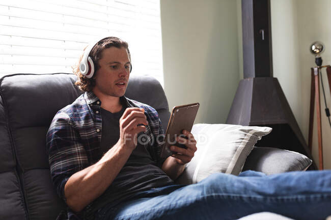 Kaukasischer behinderter Mann mit Kopfhörer und digitalem Tablet, der zu Hause auf der Couch sitzt. Behinderten- und Behindertenkonzept — Stockfoto