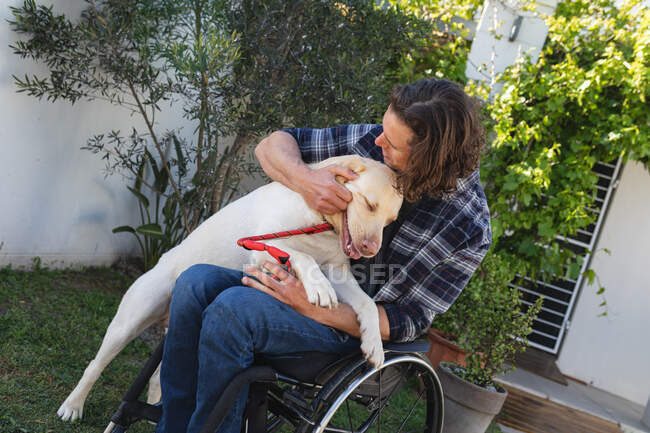 Uomo disabile caucasico seduto sulla sedia a rotelle a giocare con il suo cane in giardino. concetto di disabilità e handicap — Foto stock