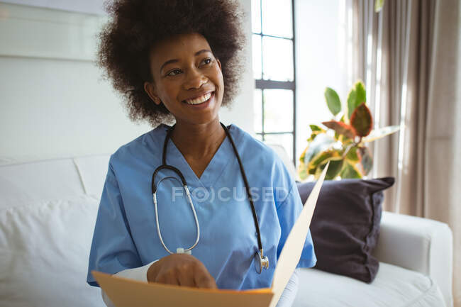 Smiling african american female doctor holding documents at home. healthcare and lifestyle during covid 19 pandemic. — Stock Photo
