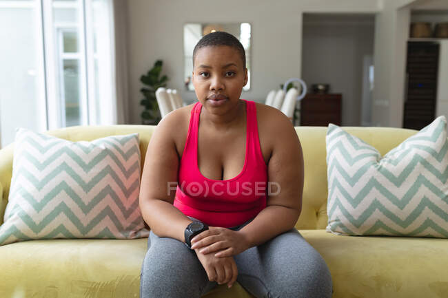 Femme afro-américaine plus la taille dans des vêtements de sport assis sur le canapé et regardant la caméra. forme physique et mode de vie sain et actif. — Photo de stock