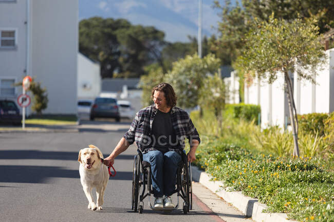 Hombre caucásico discapacitado con perro sentado en silla de ruedas en la carretera. concepto de discapacidad y discapacidad - foto de stock