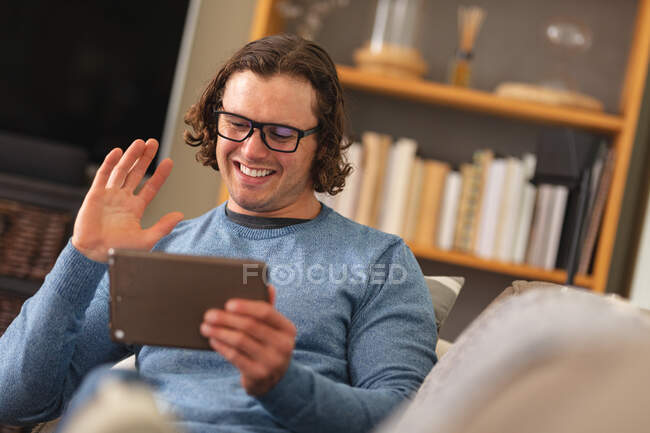 Homem deficiente caucasiano usando óculos acenando enquanto faz uma videochamada em tablet digital em casa. conceito de deficiência e deficiência — Fotografia de Stock
