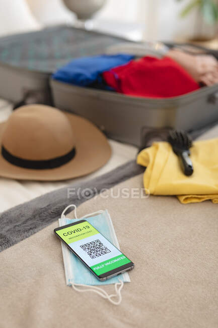 Смартфон с ковидовым паспортом, маской для лица и чемоданом, лежащим на кровати. подготовка к путешествиям во время пандемии ковида 19. — стоковое фото