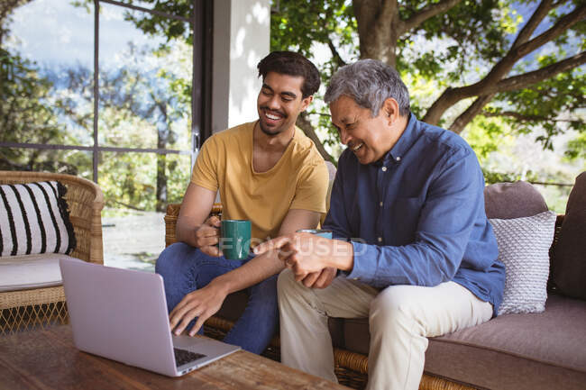 Smiling biracial adult son and senior father using laptop and drinking coffee in garden. family time at home together. — Stock Photo