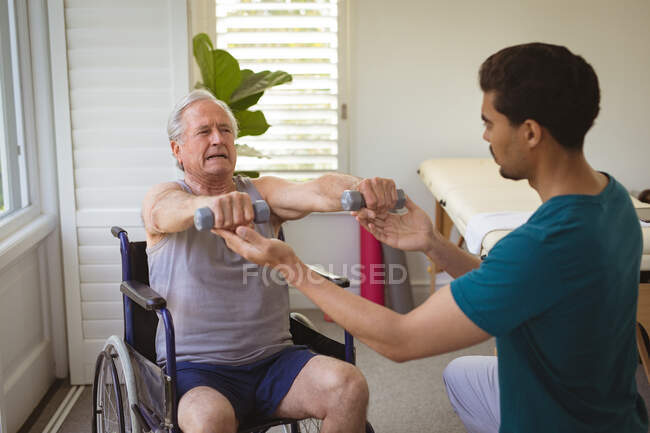 Biracial male physiotherapist treating arms of senior male patient on wheelchair at clinic. senior healthcare and medical physiotherapy treatment. — Stock Photo