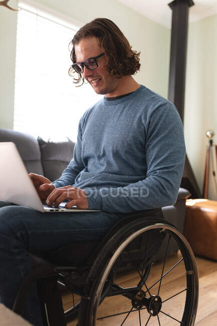 Белый инвалид в очках, сидящий на инвалидной коляске с ноутбуком дома. Концепция инвалидности и инвалидности — стоковое фото