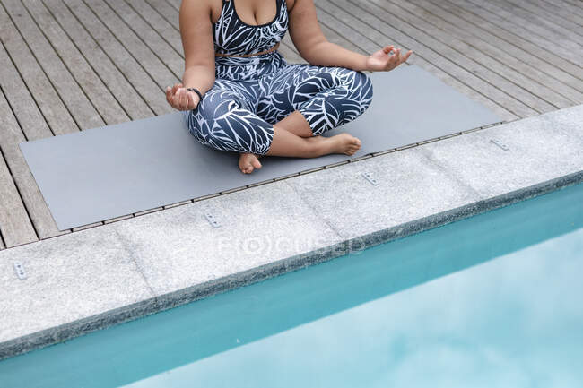 Sección media de mujer afroamericana de talla grande practicando yoga sobre estera en jardín junto a piscina. fitness y estilo de vida saludable y activo. - foto de stock