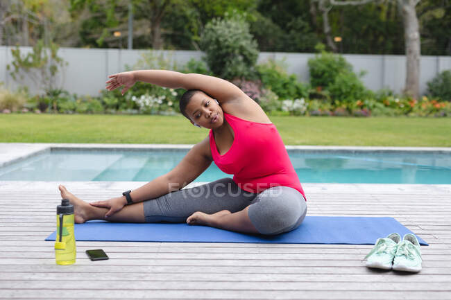 Femme afro-américaine plus la taille pratiquant le yoga dans le jardin assis près de la piscine. forme physique et mode de vie sain et actif. — Photo de stock
