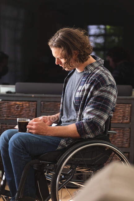 Hombre caucásico discapacitado sentado en silla de ruedas sosteniendo taza de café leyendo un libro en casa. concepto de discapacidad y discapacidad - foto de stock