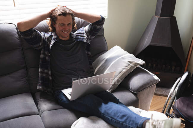 Белый инвалид, улыбающийся с помощью ноутбука, сидит дома на диване. Концепция инвалидности и инвалидности — стоковое фото