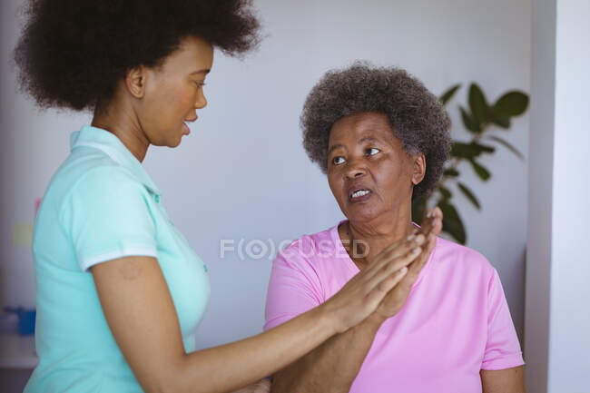 African american female physiotherapist treating arms of senior female patient and talking at clinic. senior healthcare and medical physiotherapy treatment. — Stock Photo