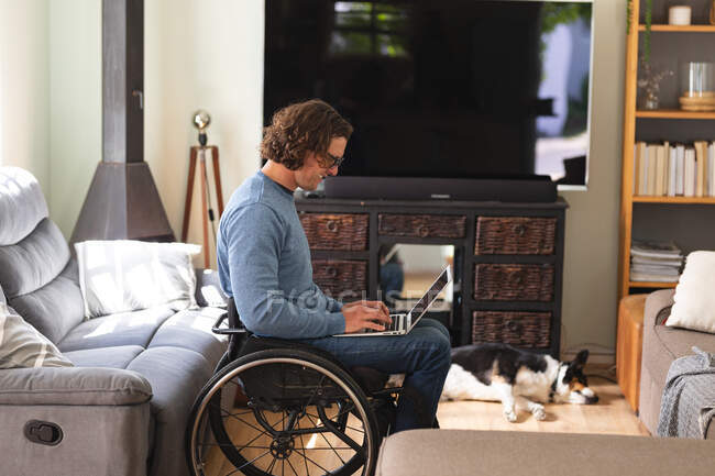 Homem deficiente caucasiano usando óculos sentados em cadeira de rodas usando laptop em casa. conceito de deficiência e deficiência — Fotografia de Stock