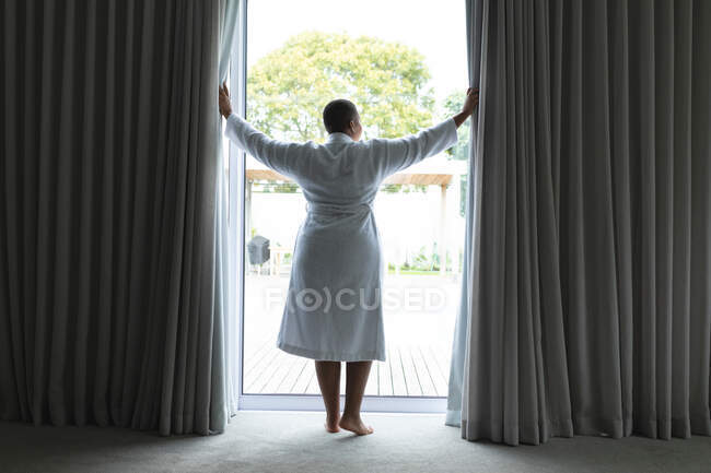 Vue arrière de la femme afro-américaine plus la taille debout à la fenêtre. mode de vie, loisirs et passer du temps à la maison. — Photo de stock