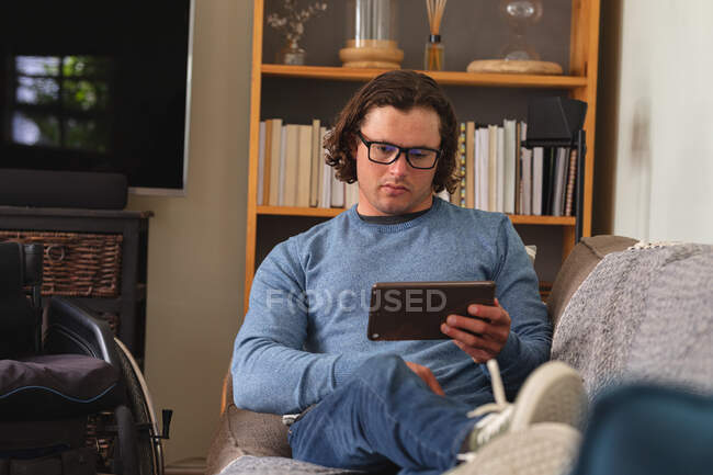 Kaukasischer Behinderter mit Brille und digitalem Tablet sitzt zu Hause auf der Couch. Behinderten- und Behindertenkonzept — Stockfoto