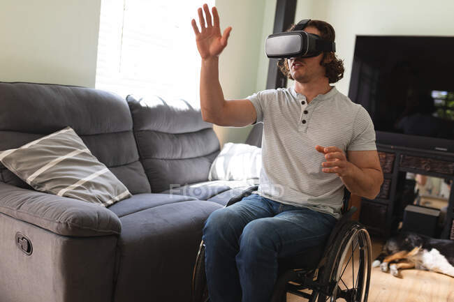 Hombre caucásico discapacitado sentado en silla de ruedas con auriculares vr y gestos en casa. concepto de discapacidad y discapacidad - foto de stock