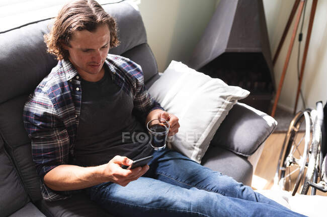 Белый инвалид держит кофейную чашку, используя смартфон, сидящий дома на диване. Концепция инвалидности и инвалидности — стоковое фото