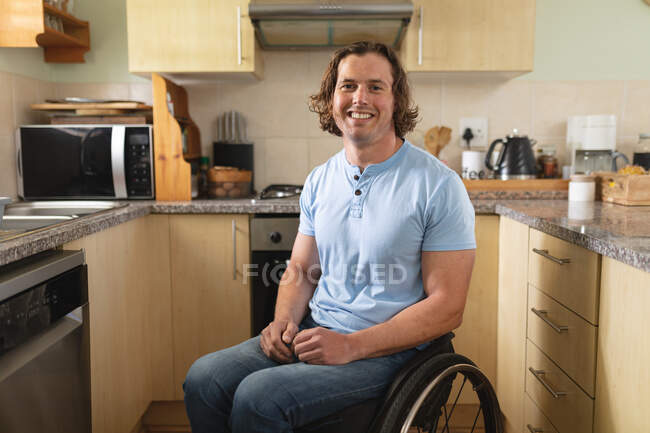 Портрет кавказского инвалида, сидящего на инвалидной коляске и улыбающегося дома на кухне. Концепция инвалидности и инвалидности — стоковое фото