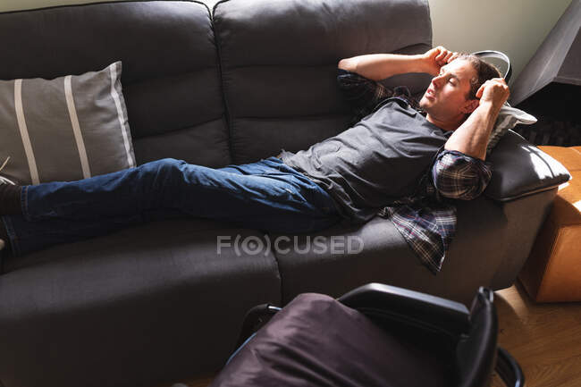 Kaukasischer Behinderter, der Kopfhörer trägt, während er zu Hause auf der Couch liegt. Behinderten- und Behindertenkonzept — Stockfoto
