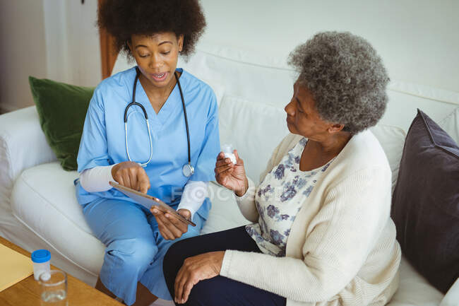 Médico afroamericano usando tableta con paciente mayor en casa. salud y estilo de vida durante la pandemia de covid 19. - foto de stock