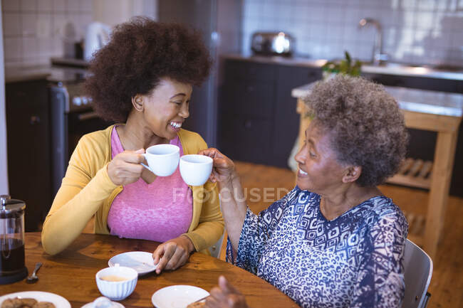 Улыбающаяся африканская старшая американка со взрослой дочерью пьет кофе. семейное время дома вместе. — стоковое фото