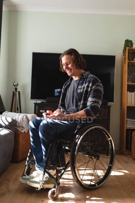 Кавказский инвалид сидит на инвалидной коляске и улыбается, пользуясь смартфоном дома. Концепция инвалидности и инвалидности — стоковое фото